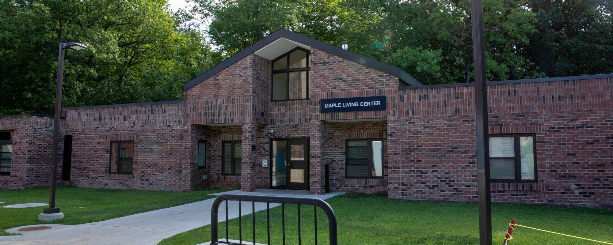A brick building with a sign "Maple Living Center" under a triangular roof. Surrounded by greenery, it conveys a welcoming, peaceful atmosphere.